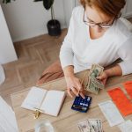 Visão de cima de uma mulher organizando contas, dinheiro e uma calculadora para controlar as despesas domésticas.