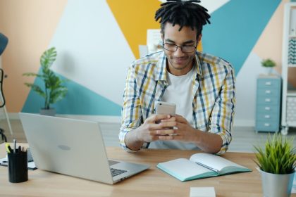 Jovem consulta o celular sentado à mesa, com um laptop ao lado, em um escritório moderno.