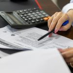Mão de uma mulher analisando documentos financeiros ao lado de uma calculadora sobre a mesa