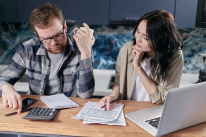 Casal analisa contas e documentos diante de um laptop, discutindo as finanças domésticas.