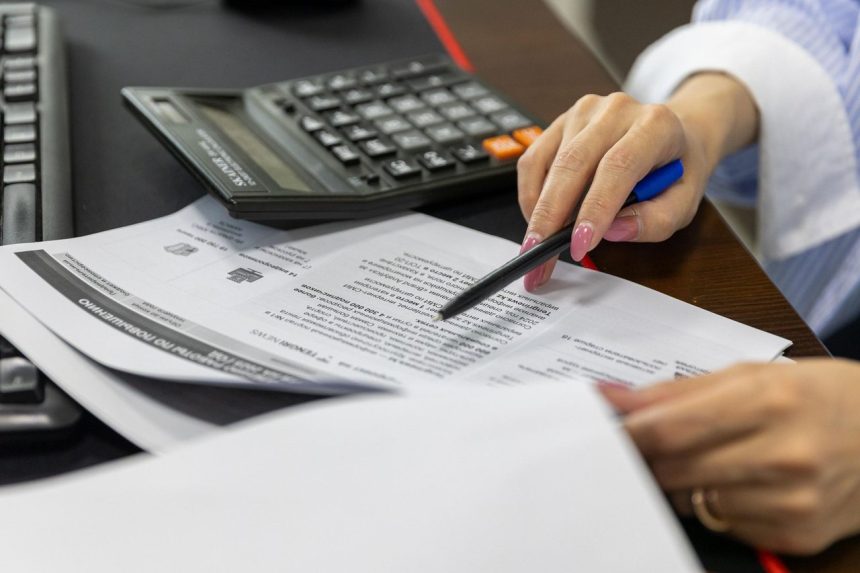Mão de uma mulher analisando documentos financeiros ao lado de uma calculadora sobre a mesa
